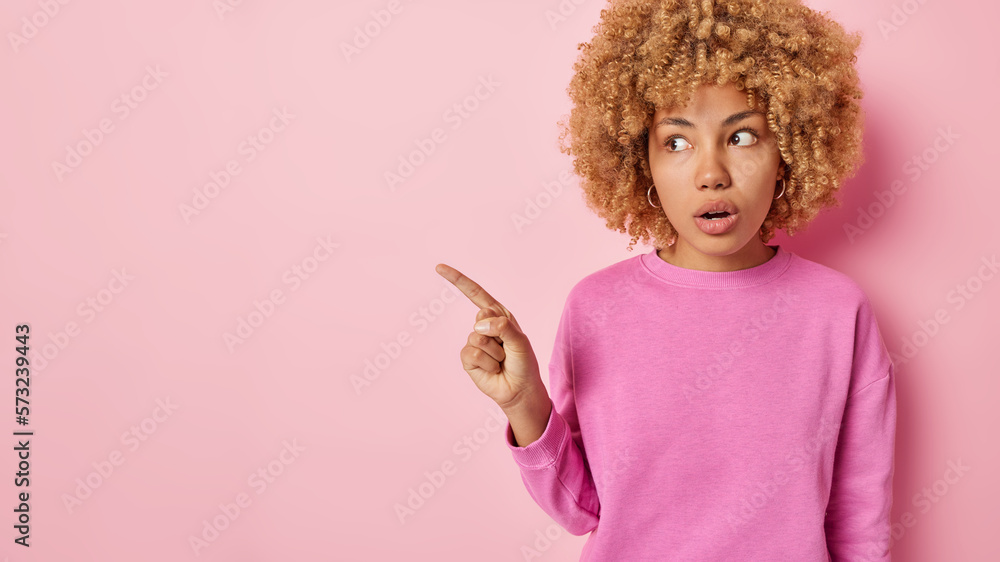 Horizontal shot of surprised curly haired woman holds breath from amazement points index finger aside on blank space wears casual jumper isolated over pink background. Omg look at this offer