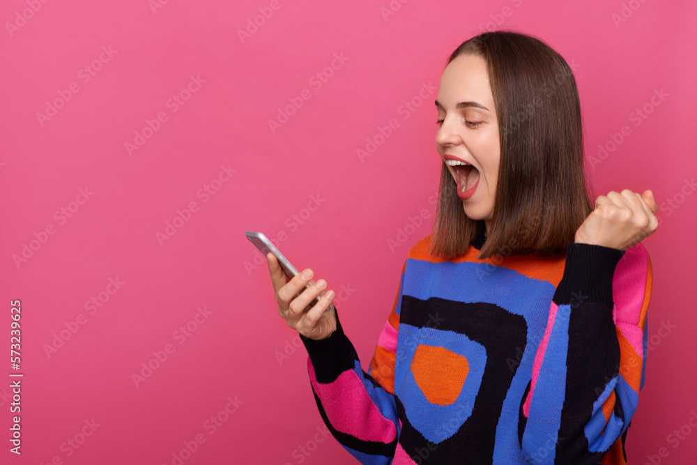 Indoor shot of extremely happy joyful woman wearing sweater posing against pink wall, using cell phone, betting and winning, rejoices, copy space for promotion. Indoor shot of extremely happy joyful woman wearing sweater posing against pink wall, using cell phone, betting and winning, rejoices, copy space for promotion.