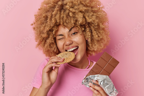 Cheerful curly haired woman bites delicious sweet cookie holds bar of chocolate breaks diet poses with unhealthy snack dressed in casual t shirt isolated over pink background. Harmful food concept