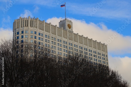The White House of the Government of the Russian Federation with the Russian flag on the roof - against a blue sky with white clouds and tree branches in the foreground.