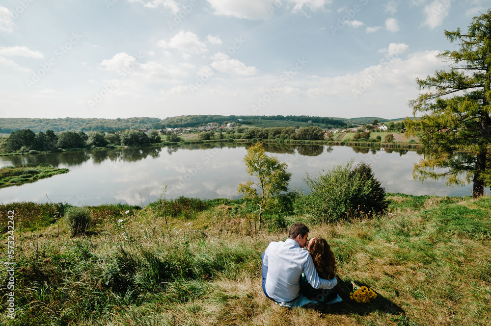 Loving couple sitting by the lake and admiring the scenery. Happy woman and man kissing on nature. Family hugging together on a sunny summer day. Back view.