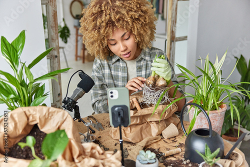 Indoor shot of female blogger botanist records video broadcasts livestream shoots content for blog holds bulb plant shoots process of replanting houseplants wears shirt and gloves. Home gardening