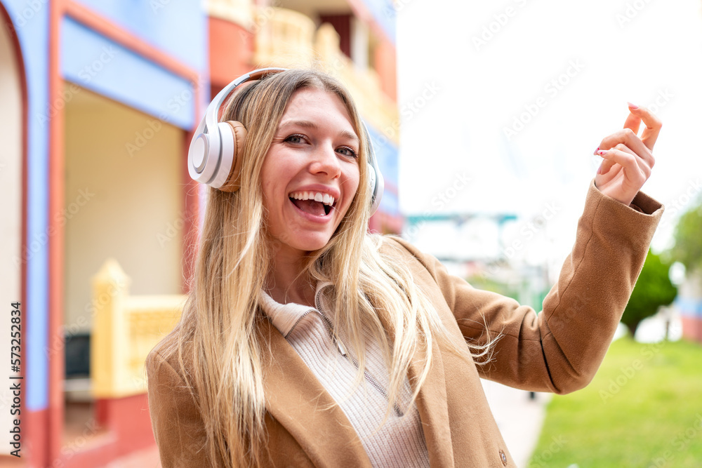 Young pretty blonde woman at outdoors listening music and doing guitar gesture