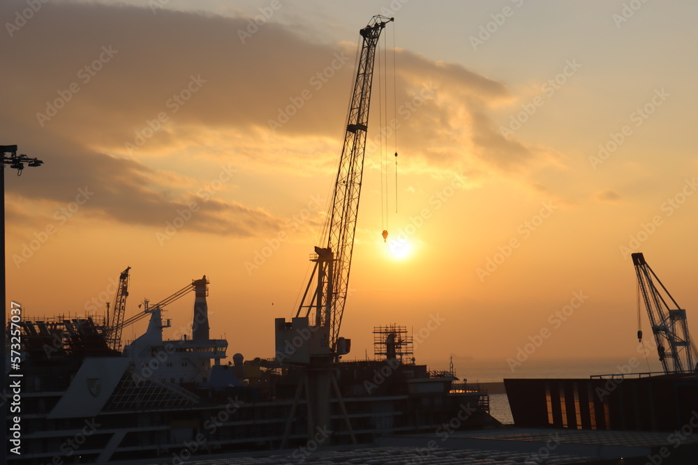 Genova, Italy - February 5, 2023: Beautiful view to the port of Genova in winter with sunset colours reflected on the water.
Golden hour hidden behind the clouds and the industrial zone of the port.