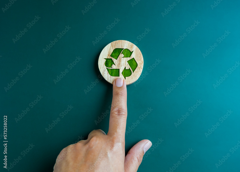 Green recycle icon symbol on round wooden block, pointing by hand isolated on blue background. Ecology, sustainability cooperation, reduce, reuse and recycle concepts. Campaign for green recycling. Green recycle icon symbol on round wooden block, pointing by hand isolated on blue background. Ecology, sustainability cooperation, reduce, reuse and recycle concepts. Campaign for green recycling.