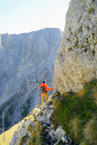 A man in a bright jacket stopped at a mountain cliff, holding a rock with his hand. Photo from the back of a man doing outdoor activities - hiking in the mountains of Montenegro