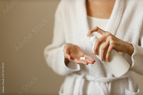 Fototapet Young woman in white housecoat pours lotion or hand moisturizer from white bottle into the palm of her hand