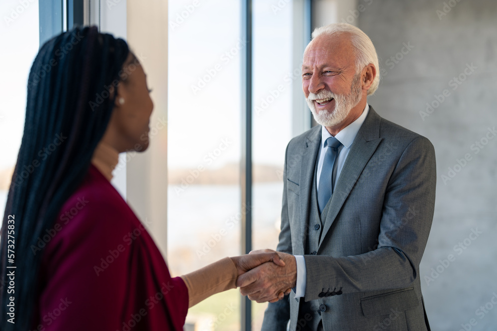 Happy confident and excited senior businessman welcoming young female employee intern on job in corporate department. Friendly senior executive shaking hand with new staff member. Happy confident and excited senior businessman welcoming young female employee intern on job in corporate department. Friendly senior executive shaking hand with new staff member.