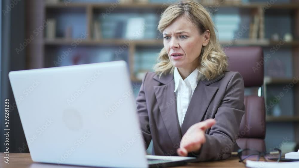 Disoriented mature businesswoman looking confusedly at laptop screen at desk at workplace in office. Sad female spreads her hands and cannot understand the mistakes in the work of the computer program