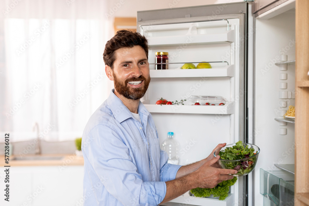 Glad hungry attractive adult caucasian man with beard takes out fresh salad from refrigerator, enjoy organic meal