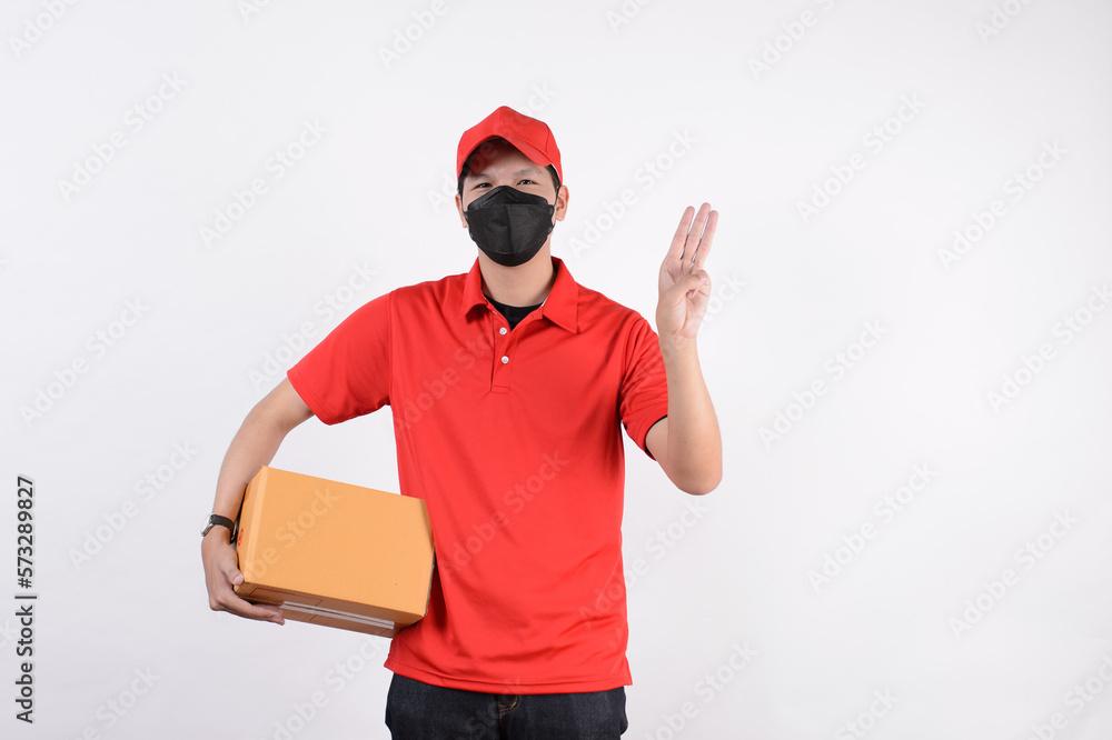 Delivery man employee in red cap blank t-shirt uniform face mask cardboard box isolated on white background. Service quarantine pandemic in front Delivery man employee in red cap blank t-shirt uniform face mask cardboard box isolated on white background. Service quarantine pandemic in front