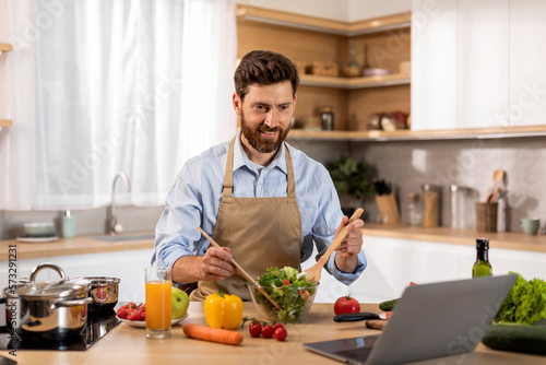 Smiling mature caucasian male in apron preparing dinner  watching video lesson on computer