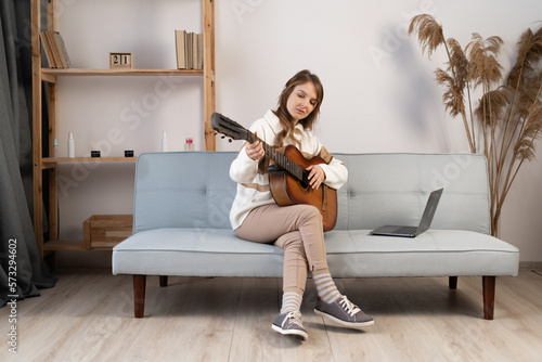 Attractive woman learning to play guitar using laptop computer sitting on sofa at home
