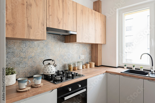 Interior of small kitchen with sink near window and wooden cabinets in kitchen furniture. Stove with gas and kettle. Modern pattern tiles on the wall.