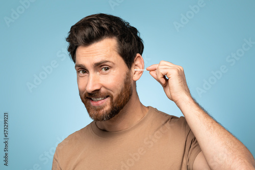 Happy handsome man using cotton swab stick for ear clean, posing over blue background and smiling at camera