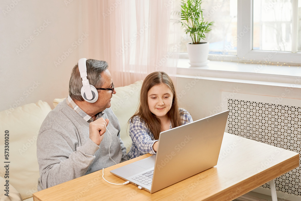 Granddaughter helping grandpa to make on line communication on laptop, listening to modern music, old man with his granddaughter using laptop making video call Granddaughter helping grandpa to make on line communication on laptop, listening to modern music, old man with his granddaughter using laptop making video call