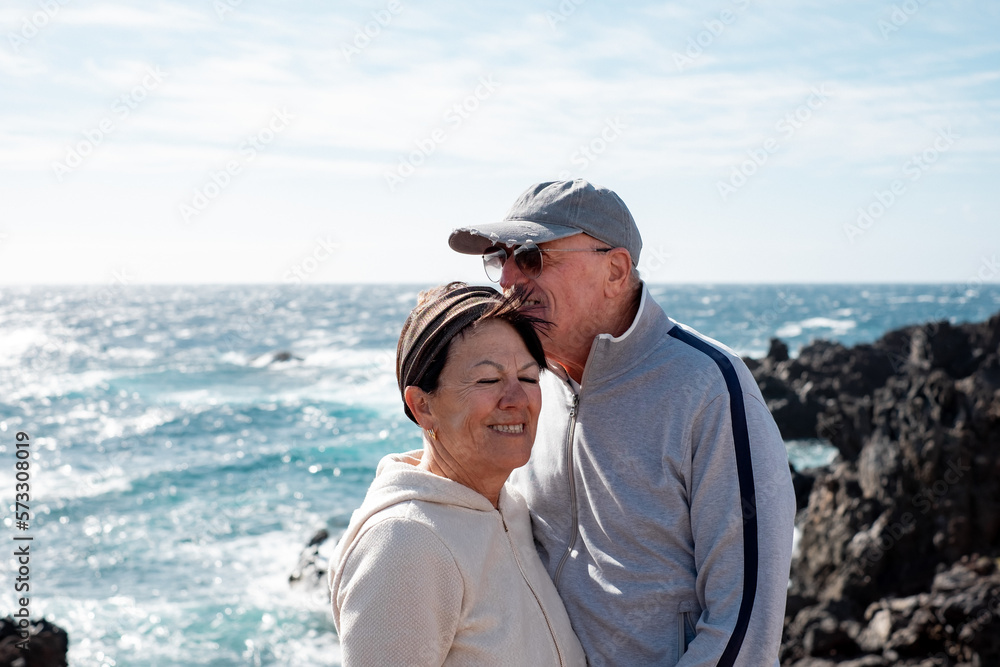Smiling caucasian senior couple standing on the cliff enjoying sunny day in travel excursion. Elderly people in vacation or retirement at sea