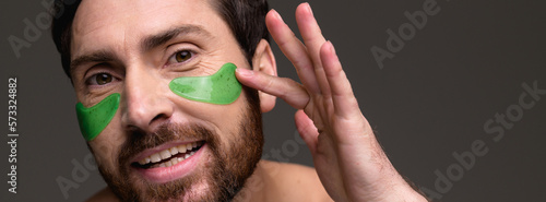 Banner view of smiling bearded man applying hydrogel eye patch isolated on grey background 
