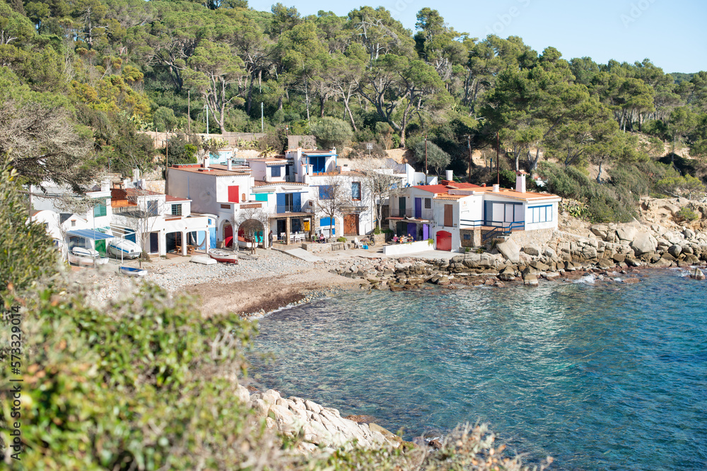 Little fisherman town with colorful huts and blue sea, surrounded with pine trees and people bask in the sun. Picturesque scene. Traveling and hiking destination - Cala s'Alguer