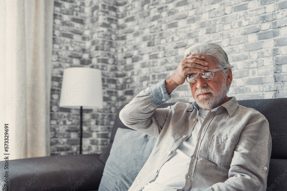 Fototapeta premium Frustrated unhealthy senior mature man touching head, having painful feelings sitting alone at home. Unhappy middle aged retired grandfather suffering from high blood pressure disease indoors..
