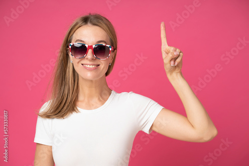 Beautiful young woman wearing casual clothes and glasses pointing finger up with successful idea. exited and happy. number one. Pretty young girl in white t-shirt gesturing up with finger on pink wall