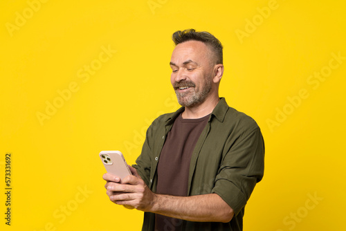 Cheerful mature stubble Man Using Mobile Application On Cellphone Or Texting Via Smartphone Communicating Online Standing In Studio Over Yellow Background. Communication And Technology for 50s