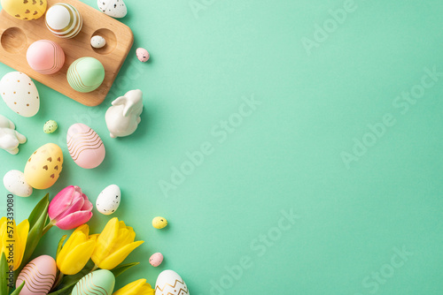 Easter decorations concept. Top view photo of bouquet of yellow and pink tulips ceramic easter bunny and colorful eggs in wooden holder on isolated turquoise background with empty space photo