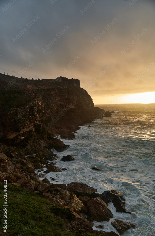 View of the sunset and the old lighthouse on a rock with a fortress on the coast of the Atlantic Ocean in the city of Nazare, Portugal. High quality photo