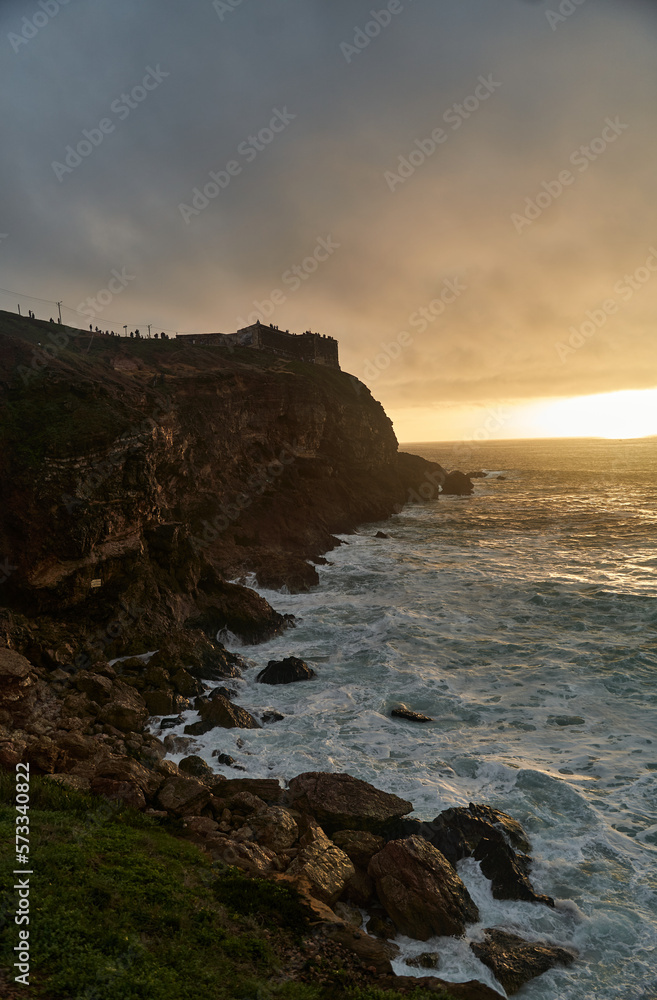 View of the sunset and the old lighthouse on a rock with a fortress on the coast of the Atlantic Ocean in the city of Nazare, Portugal. High quality photo View of the sunset and the old lighthouse on a rock with a fortress on the coast of the Atlantic Ocean in the city of Nazare, Portugal. High quality photo