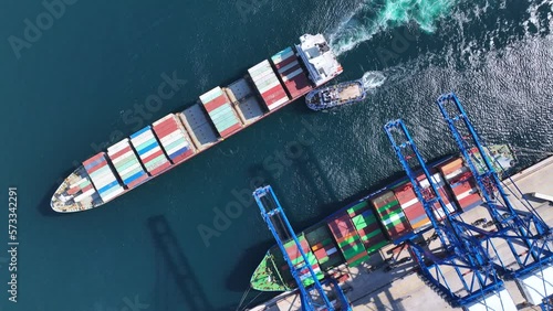 Aerial top down video of industrial cargo container ship being towed to logistics container terminal port to dock