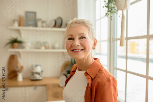 Indoor face portrait of good-looking attractive blonde lady grandmother with short hair posing against kitchen interior smiling  standing next to big window  dressed in cool stylish clothes