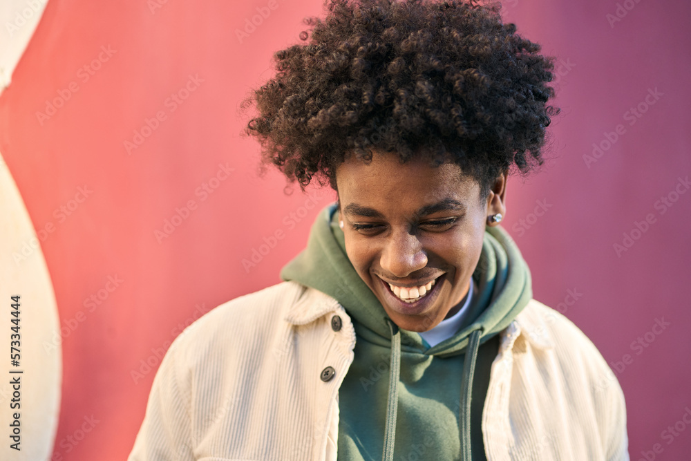 Authentic candid shot of young happy African American cool teen guy laughing on red wall lit with sunlight. Smiling cool rebel gen z teenager model standing outdoors having fun, portrait outdoors. - obrazy, fototapety, plakaty 