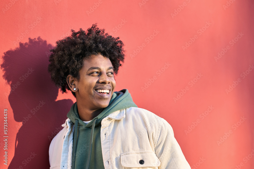 Authentic photo of young happy African American cool hipster teen guy face laughing on red city wall lit with sunlight. Smiling cool rebel gen z teenager model looking away at advertising copy space. Authentic photo of young happy African American cool hipster teen guy face laughing on red city wall lit with sunlight. Smiling cool rebel gen z teenager model looking away at advertising copy space.