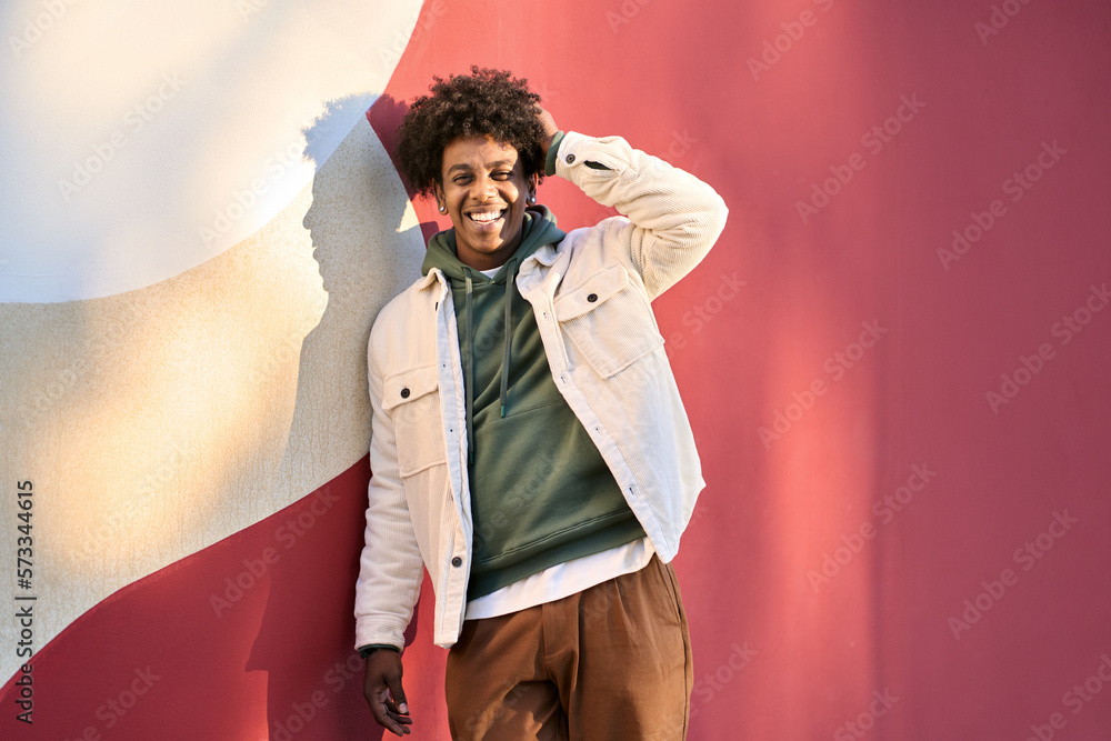 © insta_photos - Young happy African American teen guy laughing on red wall lit with sunlight. Smiling cool ethnic generation z teenager student model standing looking at camera posing for portrait outdoors.
