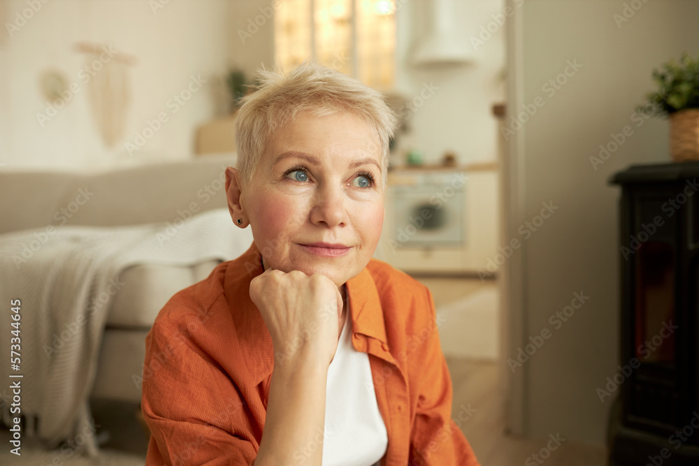 Portrait of beautiful senior woman with short stylish blond hair sitting on floor in living room, contemplating, reflecting, recollecting best memories of her life, feeling nostalgia about past Portrait of beautiful senior woman with short stylish blond hair sitting on floor in living room, contemplating, reflecting, recollecting best memories of her life, feeling nostalgia about past