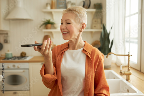 Portrait of pretty retired female with stylish haircut recording voice message on smartphone communicating with friends of group chat in popular messenger standing in stylish cozy kitchen