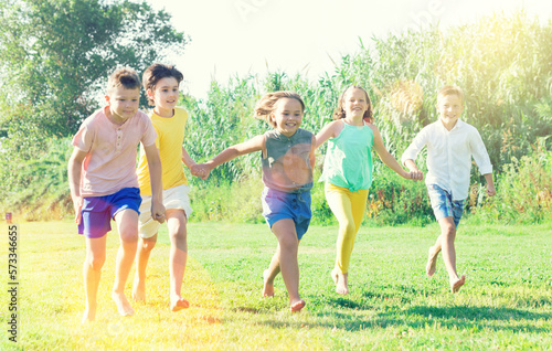 Portrait of cheerful preteen boys and girls running on green lawn in city park on summer day ..