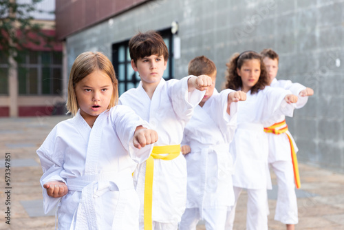 Group of kids in white kimono training outdoors on street.