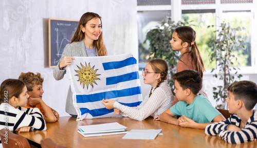 Decent teacher showing Uruguay flag to group of preteen schoolchildren in classroom during lesson