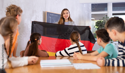 Group of curious preteen learners and smiling young female teacher with flag of Germany in classroom of academy