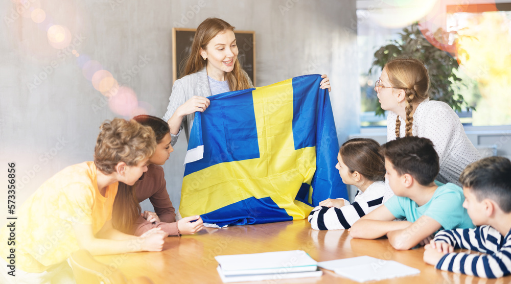 Female teacher tells her classmates about the country of Sweden, holding a flag in her hands