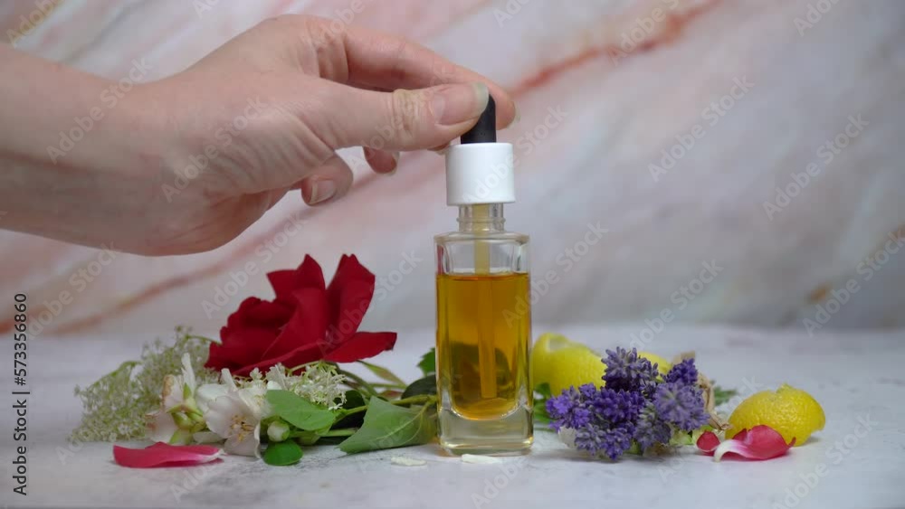 A woman uses natural face serum in a transparent dropper bottle with lavender, rose, lemon, jasmine and elderflowers on marble background. A woman uses natural face serum in a transparent dropper bottle with lavender, rose, lemon, jasmine and elderflowers on marble background.