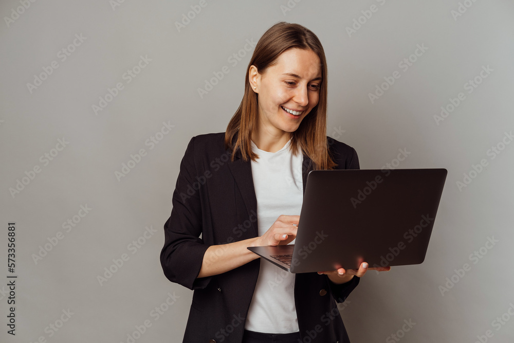 POrtrait of a cheerful smart woman is using laptop while holding one in a grey studio. POrtrait of a cheerful smart woman is using laptop while holding one in a grey studio.