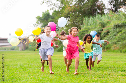 Young joyful boys and girls running through field with balloons in hands.