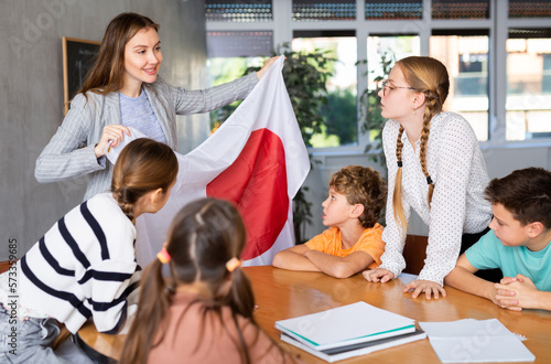 Decent teacher showing Japan flag to group of preteen schoolchildren in classroom during lesson