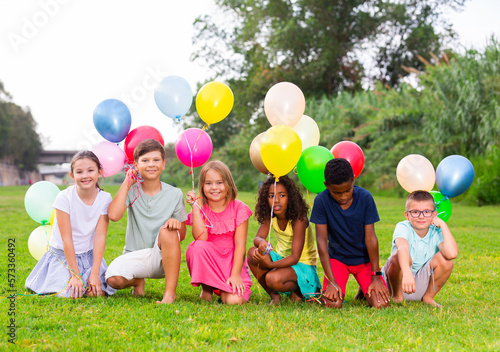 Multiracial group of cheerful tweenagers holding colored helium balloons in hands, posing together in summer city park. Happy childhood concept