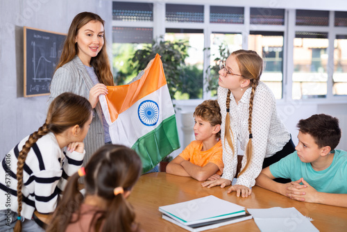 Smiling oung female teacher showing flag of India to schoolchildren preteens during history lesson in classroom