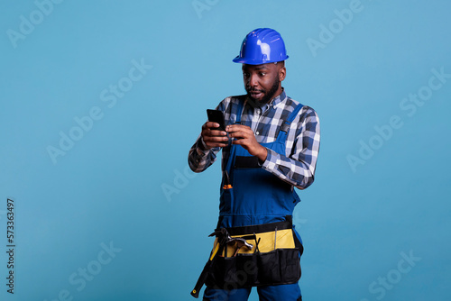 African american builder looking with amazed expression at cell phone screen, wearing coveralls and hard hat. Electrician with tool belt using mobile device against light blue background. © DC Studio