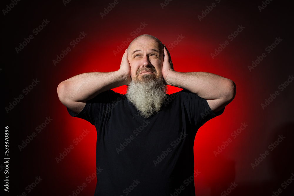 The gray-haired man covered his ears with his palms and does not want to hear anything against the background of a red spot The gray-haired man covered his ears with his palms and does not want to hear anything against the background of a red spot
