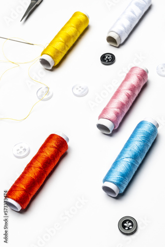 View of black and white buttons, needle with threaded yellow thread, various colors on thread spools on white background